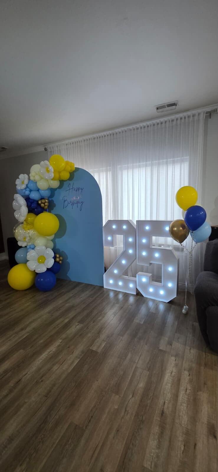 Powder-blue arch backdrop with a yellow, white and navy balloon garland, daisies and light-up '25' numbers for a 25th birthday